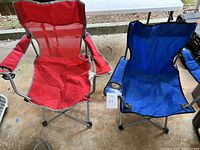 Pair of folding camp chairs side by side, red and blue, showing armrests, cup holders, and metal frames.