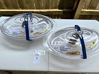 Photo showing both clear acrylic serving trays placed side by side, each with an oval shape and a secure lid with a handle, secured by blue straps.