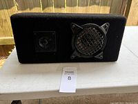 Front view of rectangular black fabric-covered speaker showing woofer with metal grill on right and tweeter on left