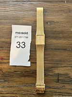Photo of gold-tone wristwatch with mesh band and square face laying flat on wooden surface.