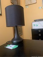 Photo of black desk lamp with a sleek, rounded base and a cylindrical black fabric lampshade placed on a dark wood surface. The lamp has a power cord connected at the base.