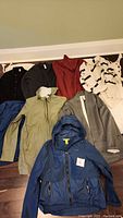 All seven clothing items arranged on floor including rain jackets, sweatshirts, shirts, cardigan