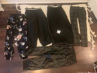 Five pairs of pants laid on a rug including floral leggings, black pants, plaid pants, and faux leather joggers