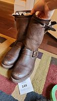 Front view of brown leather Frye boots showing rounded toe, buckle strap, and sheepskin-like lining inside.