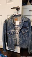 Full front view of blue denim jacket hung on hanger showing button front closure, collar, front pockets, and embroidered Les Miserables Canada text on front left pocket area.