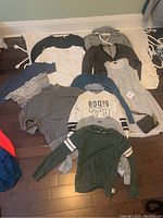 Top view of laid out clothing items including sweaters, long sleeve shirts, hooded top, and a dress or long shirt, also small black pouch.