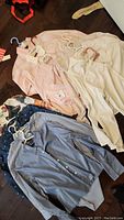 Photo showing five light sweaters and shirts in pink, cream, and striped patterns on hangers, spread on floor.
