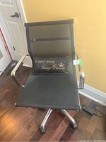 Front angle showing black mesh office chair, chrome armrests and base, and black pillow on seat with gold embroidery reading 'It ain't easy being Queen'. Chair on hardwood floor with yellow wall background.