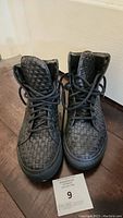 Front view of black woven high-top sneakers with laces tied, showing woven texture and shape.