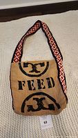 Front view of the tote bag showing the burlap texture, black printed FEED logo, and colorful geometric patterned shoulder strap.
