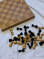 Wooden chessboard with light and dark squares, foldable with hinge, and numerous black and blonde wooden chess pieces scattered on a cloth.