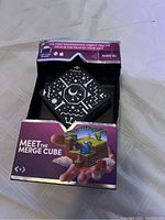 Photo showing the Merge Cube in its original packaging with visible geometric white patterns on the black cube and partial packaging text mentioning holographic object in the palm of your hand.