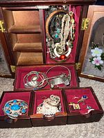 Open jewelry box showing upper compartment with hanging jewelry on twirling holder and multiple drawers with rings, bracelets, and pearl strands.
