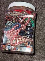Front view of the plastic jar packed with various colorful beaded necklaces and bracelets, including pink, blue, green, and pearl-like beads.