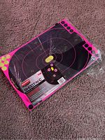 Two images showing packaged Birchwood Shoot-N-C shooting target boards with pink outline and yellow aiming dots, on a carpeted floor.