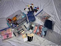 Overview of multiple sewing materials and tools organized in plastic containers along with various fabric pieces and accessories scattered on a white sheet.