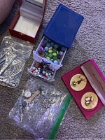 Photo of assorted jewelry showing plastic boxes with colorful beads, boxed cameo jewelry, watches in plastic bags, and silver-tone bracelets on carpeted floor