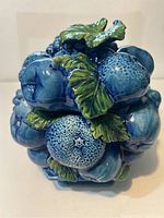 Front angle showing detailed 3D indigo blue ceramic fruit motif with green leaves on lid and side.