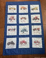 Full view of quilt showing 12 embroidered antique cars in white squares with blue border on wooden floor.
