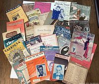 Full assortment of over 30 antique sheet music covers laid out showing various titles and colorful vintage cover art, illustrating the breadth and age of the music collection.