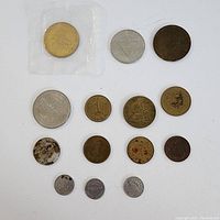 Fifteen assorted tokens and coins laid out on a white background, with various sizes and colors, some worn, one coin in sealed plastic packaging.