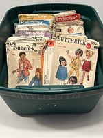 Plastic bin filled with over 30 vintage sewing pattern envelopes from brands like Butterick and Simplicity. Various styles for women and children are shown.