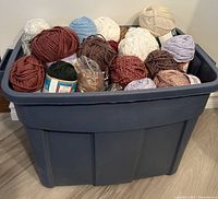 Large plastic bin filled with multiple balls and skeins of yarn in assorted colors including white, brown, blue, red, purple, tan, and green.