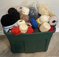 Green plastic bin filled to the top with assorted yarn skeins and cones in various colors including black, white, red, and blue.