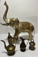 Photo showing large brass elephant figurine with raised trunk and three additional smaller brass decorative items arranged in front of it.