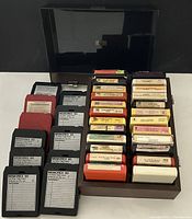 Photo showing multiple 8 track tapes arranged in rows inside a black plastic covered case, some tapes loose to the side.