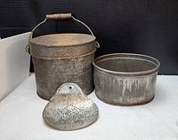 Three vintage galvanized metal items placed on a surface against a black backdrop. Larger pot has a round lid with wood handle and wire handle. Smaller open pot next to it. Metal sifter with holes in front.