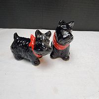 Pair of black ceramic Scottish Terrier salt and pepper shakers with red scarves, shown standing on a white surface against a dark background.