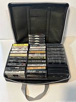 Black carrying case open showing 30 vintage cassette tapes arranged inside, visible titles on tape cases.