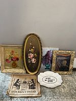 Six assorted art pieces displayed on floor against wall including framed floral paintings, an art piece resembling 'The Scream', hand-stitched fabric, and a ceramic plate with inscription.