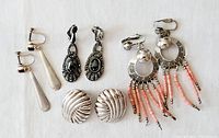 Four pairs of clip-on earrings arranged on white background: long teardrop silver pair with garnet stones, black ornate filigree pair with marcasite, round twisted silver pair, Greek key design earrings with coral stone fringe