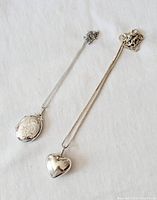 Two sterling silver pendant necklaces laid on a white surface. Left pendant is oval-shaped with floral engraving; right pendant is a smooth puffy heart shape.