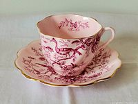 Side view of the pink on pink floral and bird design teacup resting on matching saucer, showing gold trim and scalloped edges