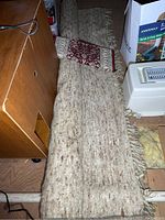 Full view of the long handmade wool area rug with neutral tones and visible tassels at the edge, placed in an attic space among other stored items.
