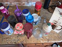 Various sized containers, beverage holders, jars, small glass vase, and thermos arranged on counter.
