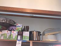 Top kitchen shelf showing very large pot with lid, smaller pot, Tupperware bakery containers, and boxes of light bulbs