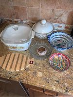 Overview of all items in the lot placed on kitchen counter showing Corning Ware casserole, Covington Edition serving dish, wooden fish trivet, glass tea light holder, glass bowl, ceramic bowl, and small decorative dish.