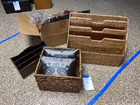 Photo showing a large rectangular wicker file storage organizer with three stacked compartments, a small black wooden three-slot letter holder, a woven wicker storage basket containing two square damask throw pillows with peacock design.