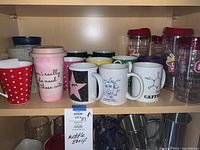 Multiple ceramic mugs, plastic covered drink dispensers, and plastic tumblers on a shelf.