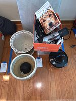 Photo showing grey ironing board with some stains, next to box of hair care accessories, book 'A Dog's Purpose', ceramic flower pots and black ceramic pumpkin decoration on floor.