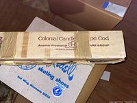 Box of Colonial Candle wedding candle and a box of Holiday brand skating shoes shown stacked on floor