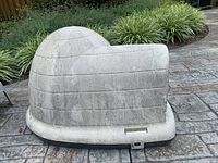 Cream colored hard plastic cat hut with igloo shape and weathering on surface placed outdoors on stone patio near greenery.