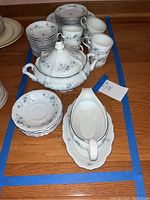 Photo of assorted blue and white china set including plates, coffee cups, a two-handled covered serving dish, and a gravy boat on a wooden table.