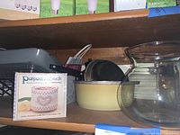 Shelf holding a Rival mini potpourri crockpot in box, metal roasting pan, yellow crepe pan, and two large clear glass vases