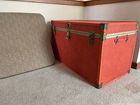 Orange vintage trunk with metal corner protectors and nail head trim, shown closed against a wall.