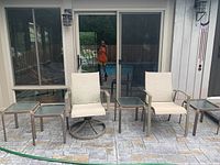 Front view of two cream fabric patio chairs, one swivel, alongside four small metal and glass top side tables on outdoor patio with stone tile flooring.